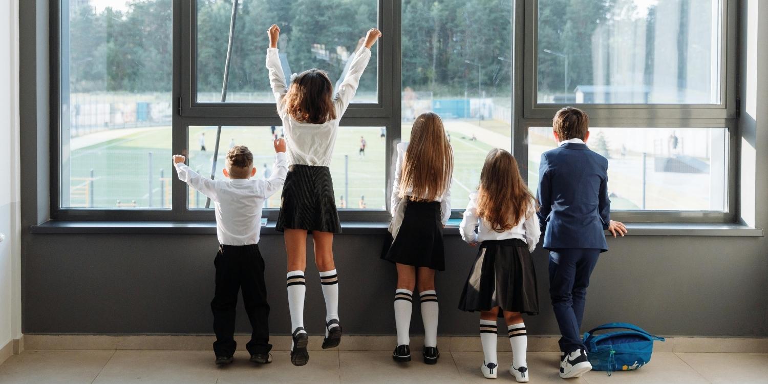 Five school children in uniforms viewed from behind, looking out large windows onto a school sports field