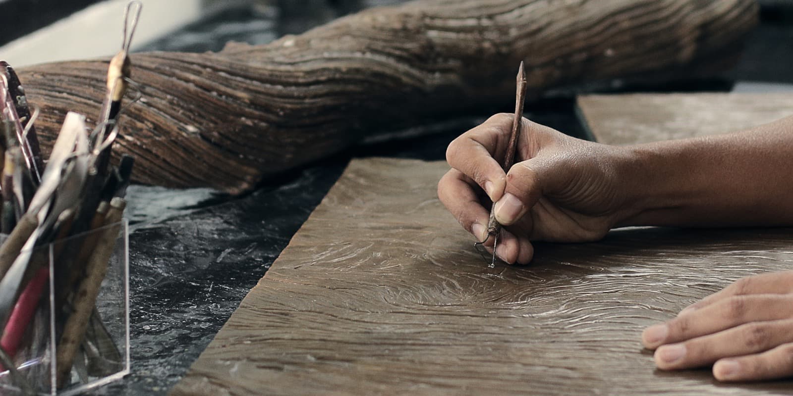 A maker incising a pattern into a clay panel with a hand tool at a workshop bench