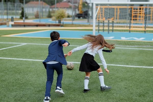 Deux enfants en uniforme scolaire jouant au football sur un terrain sportif scolaire en plein air.